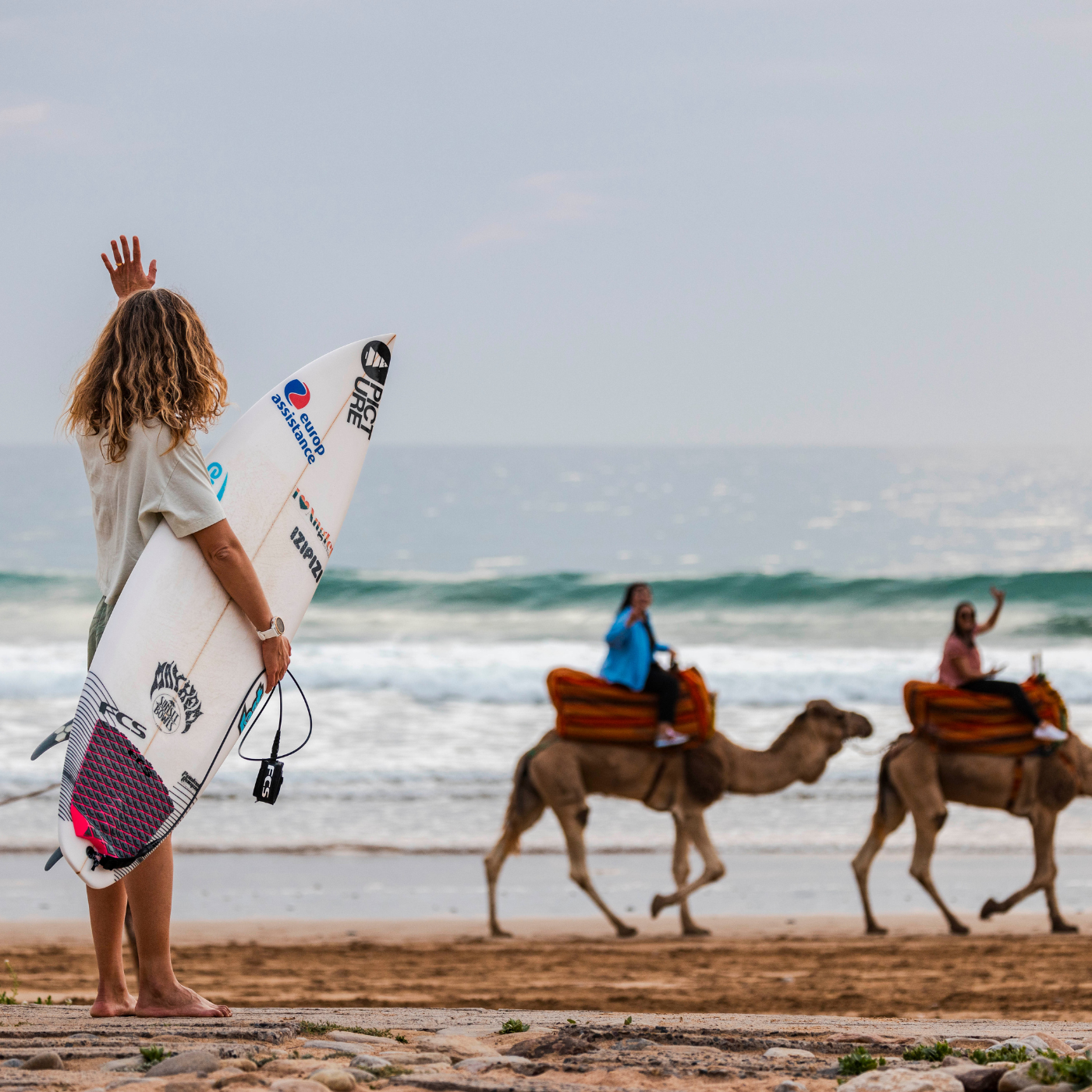 A girl surfing at Paradis Plage, waving to camels on the beach