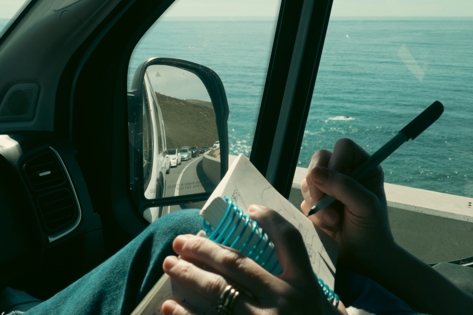 An artist sketching in a notebook in the passenger seat of a van parked above a blue ocean.