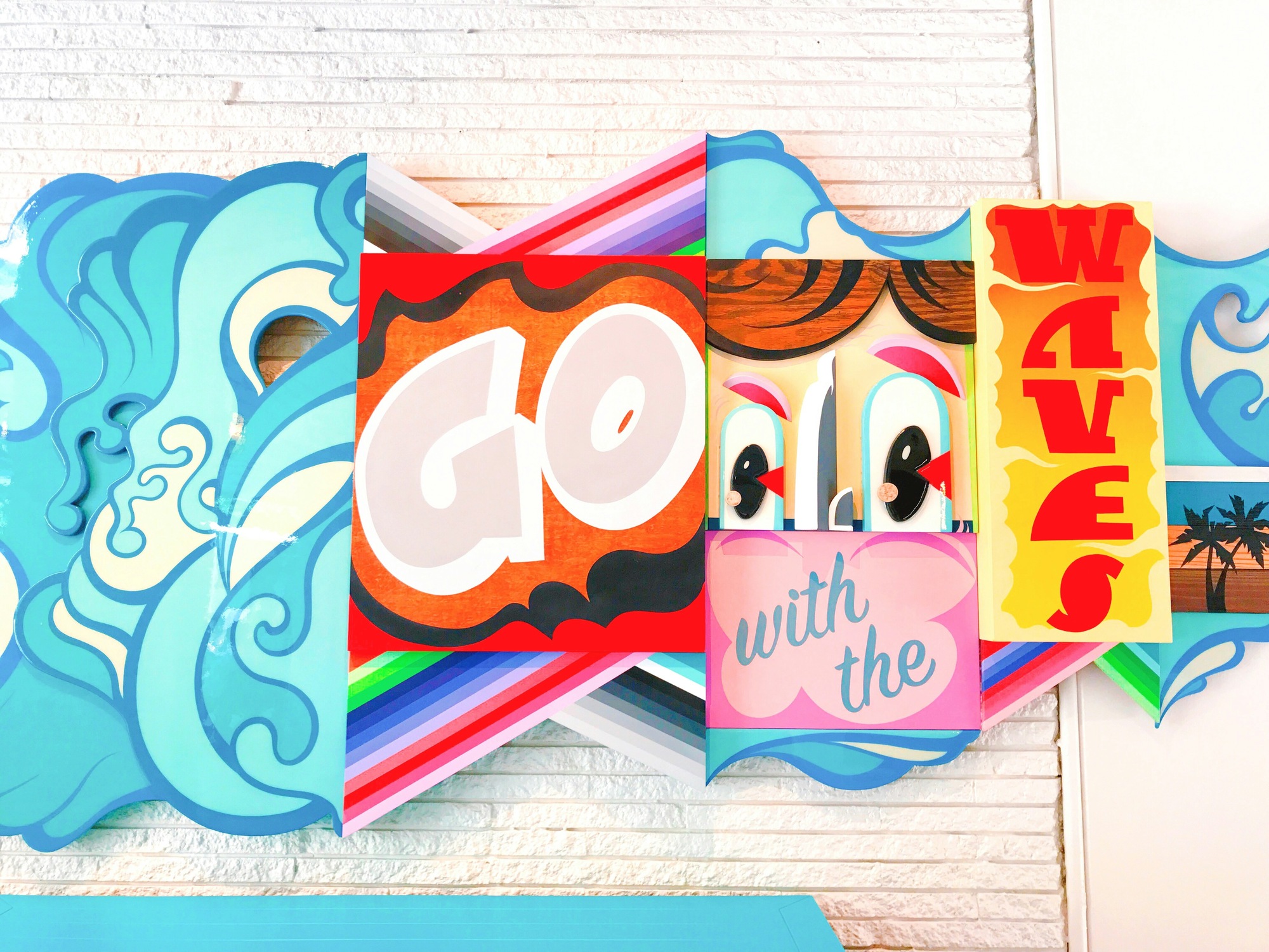 A bright pop-art wall installation of layered wave shapes and lettering reading Go With The Waves.