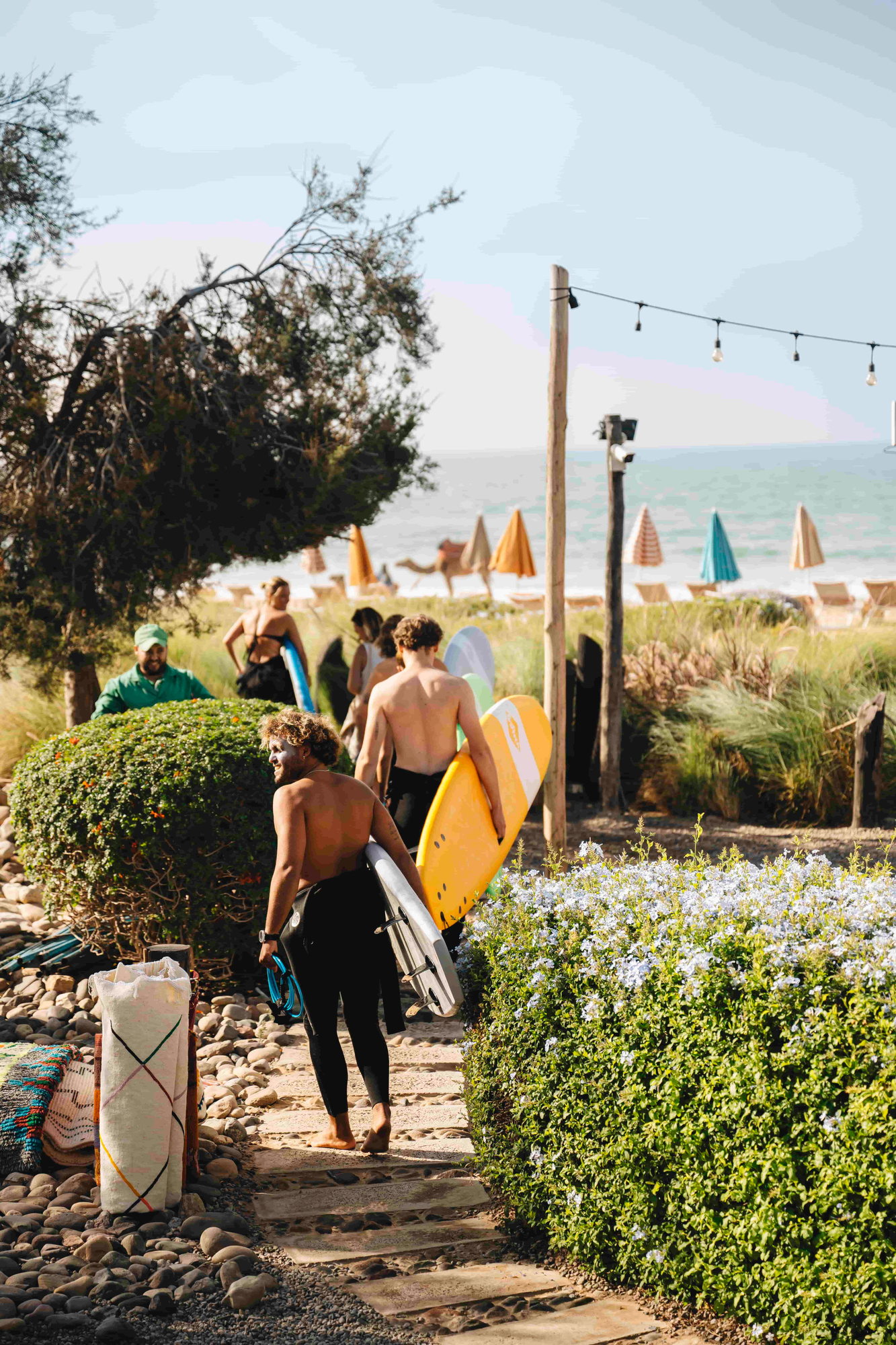 Surfers carrying boards down the beach path at Paradis Plage, Imi Ouaddar, with the Atlantic and beach parasols beyond
