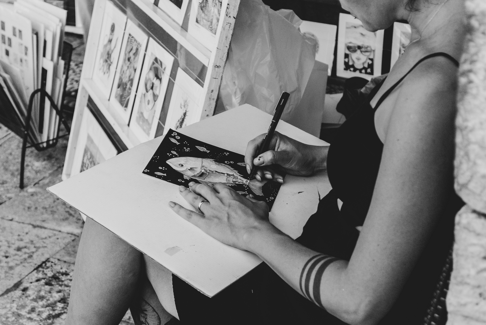 Black and white photograph of an artist inking a fish illustration on her lap at a market stall, her prints displayed behind her.