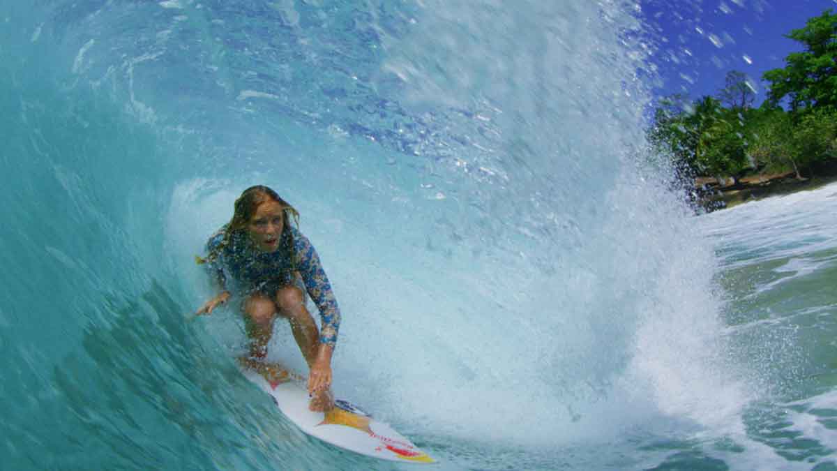 Caity Simmers crouches deep inside a turquoise barrel in a blue floral long-sleeve suit, spray blowing past her