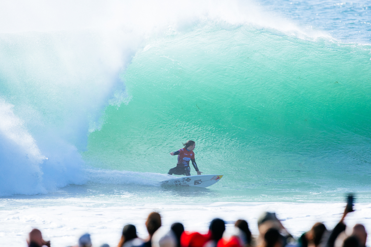 Caitlin Simmers in a red jersey crouched low inside a large turquoise barrel at Supertubos, with spectators in the foreground