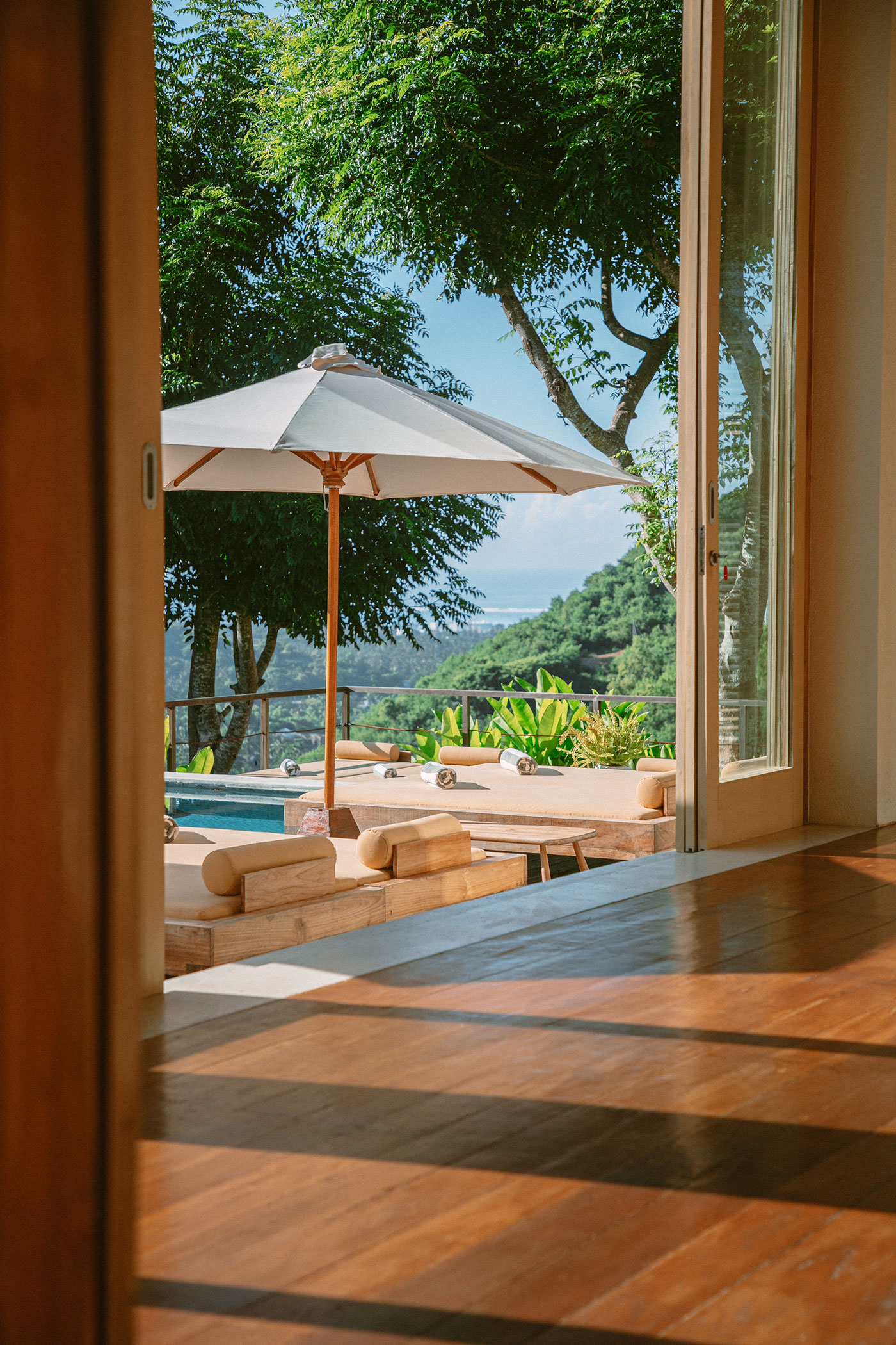 View through open glass doors to a pool deck with a cream parasol and cushioned loungers, forested hills sloping down to the sea beyond