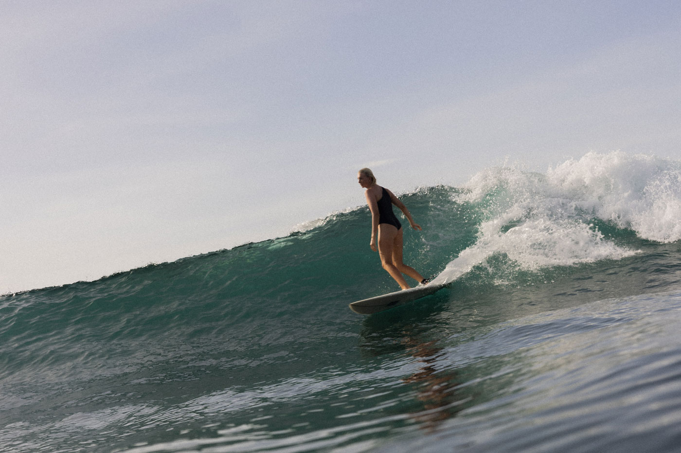 A woman in a black swimsuit trims along the green face of a breaking wave on a longboard