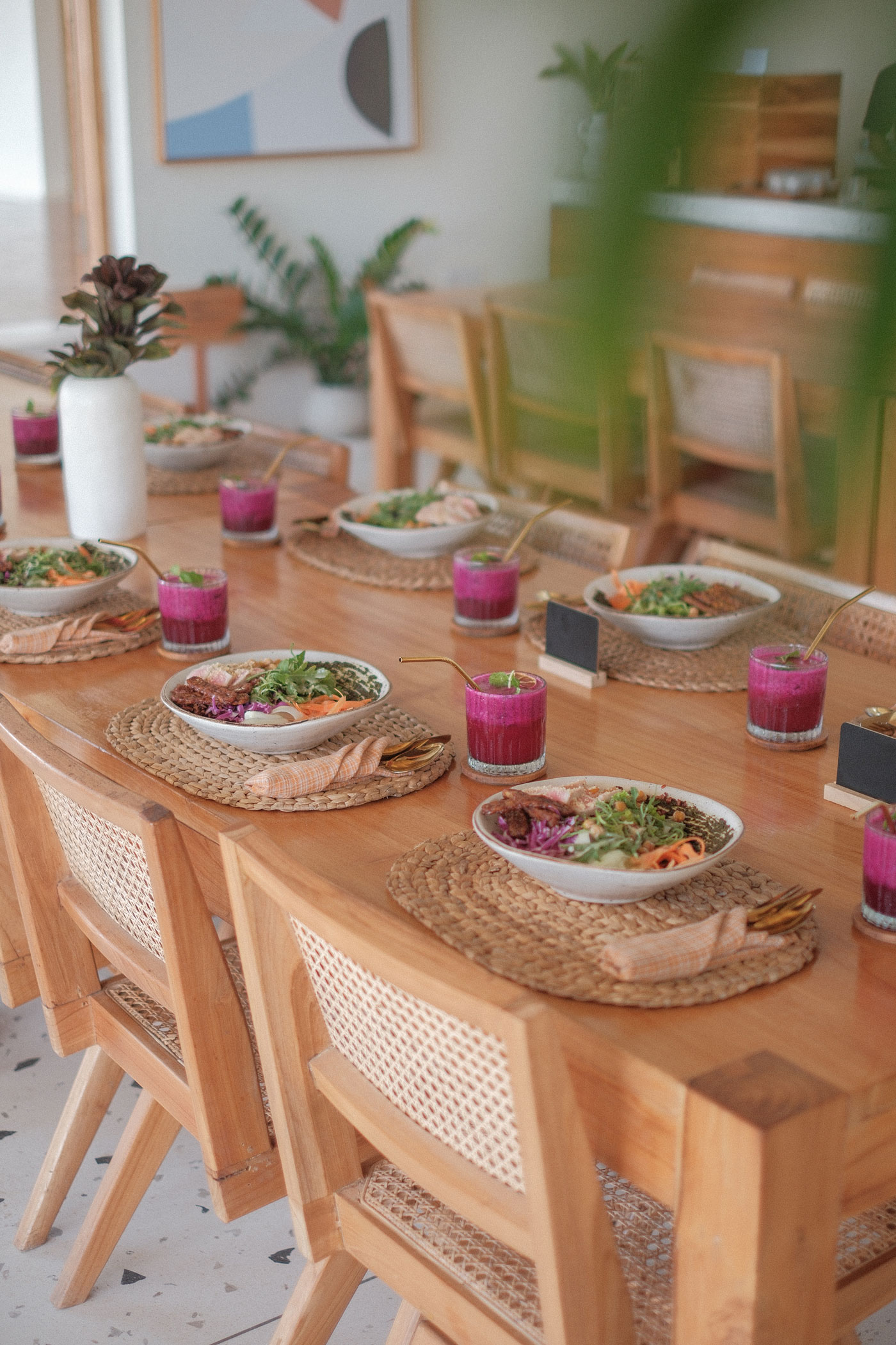 A long wooden table laid with salad bowls and pink dragon fruit drinks on woven placemats, rattan chairs alongside