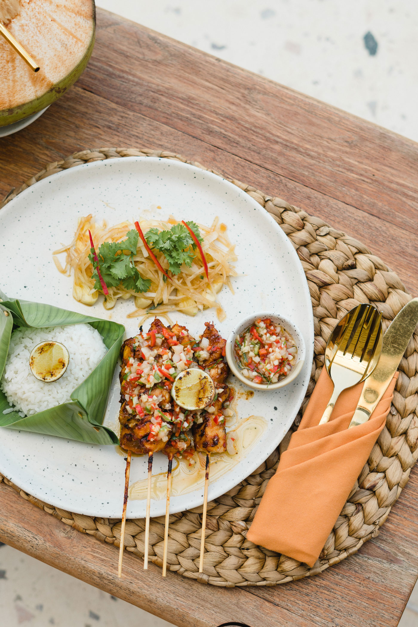 Chicken satay skewers with sambal, papaya salad and rice in a banana leaf on a speckled plate, a fresh coconut beside it