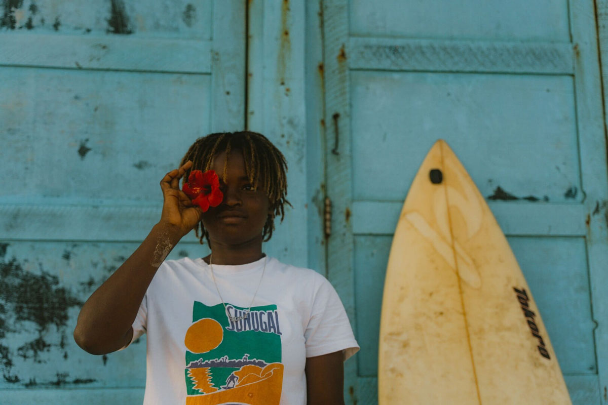 A young man holds a red flower over one eye, standing beside a surfboard against a weathered turquoise wall