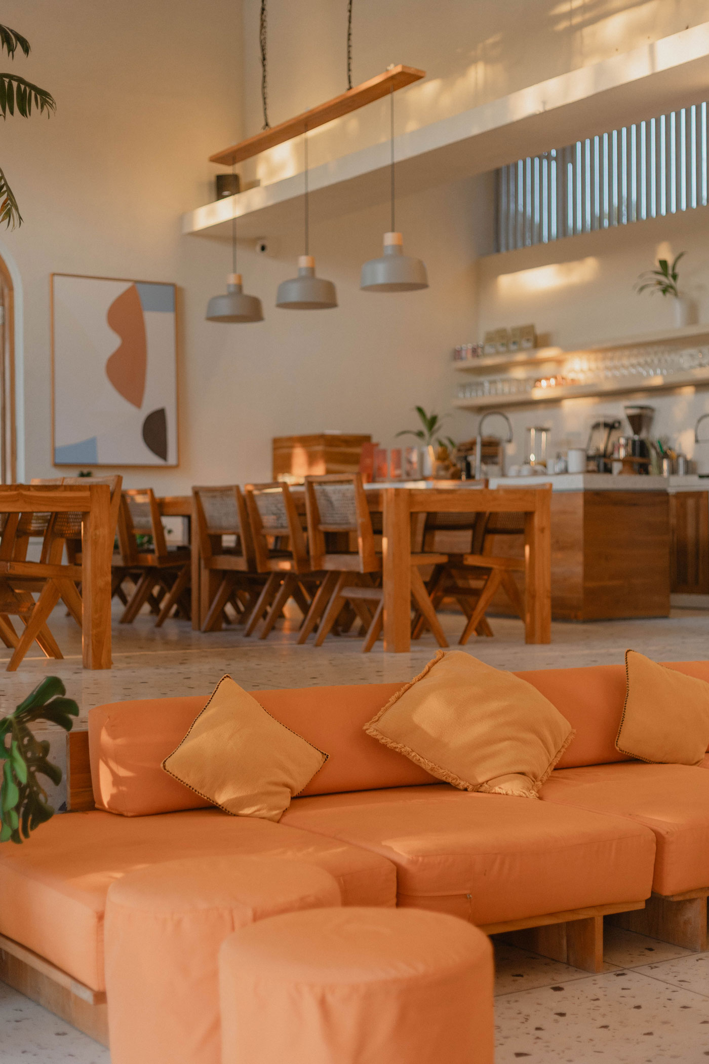 An orange sofa with mustard cushions in a high-ceilinged lounge, wooden dining tables and a coffee bar behind in evening light