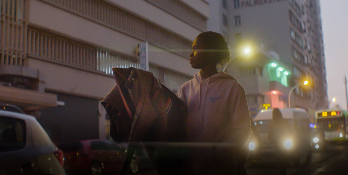 A young person in a hooded top carries a board along a city street at dusk, with traffic lights behind