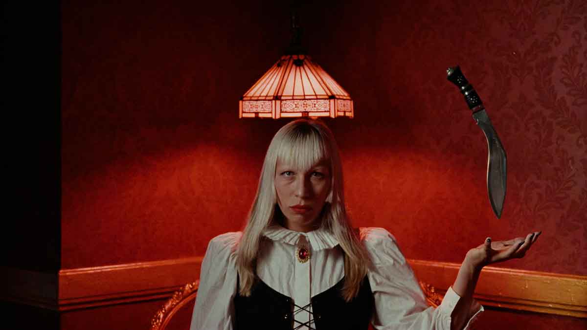 Jaleesa Vincent sits beneath a stained-glass lamp in a red wallpapered room, a curved knife hanging in the air above her open hand