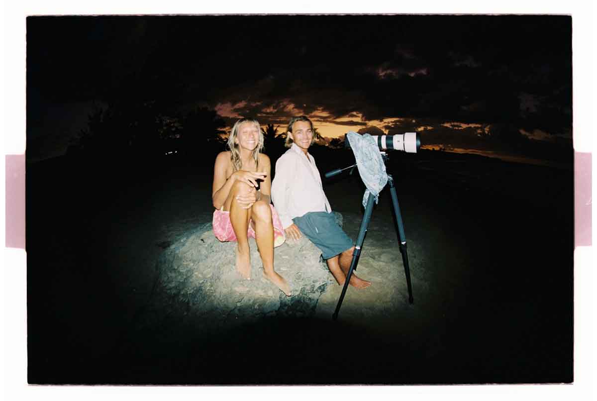 Jaleesa Vincent and Luka Raubenheimer sit on a rock at dusk, lit by a flash, beside a long-lens camera on a tripod