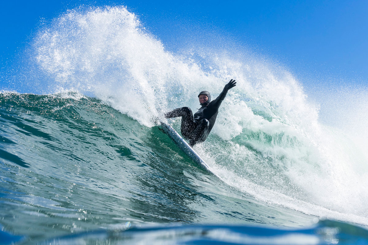 A surfer in a hooded wetsuit and gloves carves across a blue cold-water wave