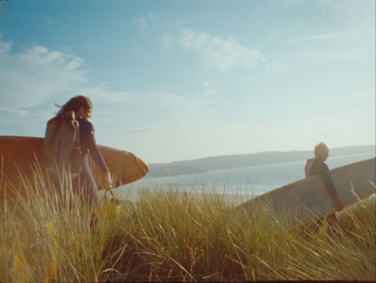 Two surfers carry their boards through tall dune grass towards the sea