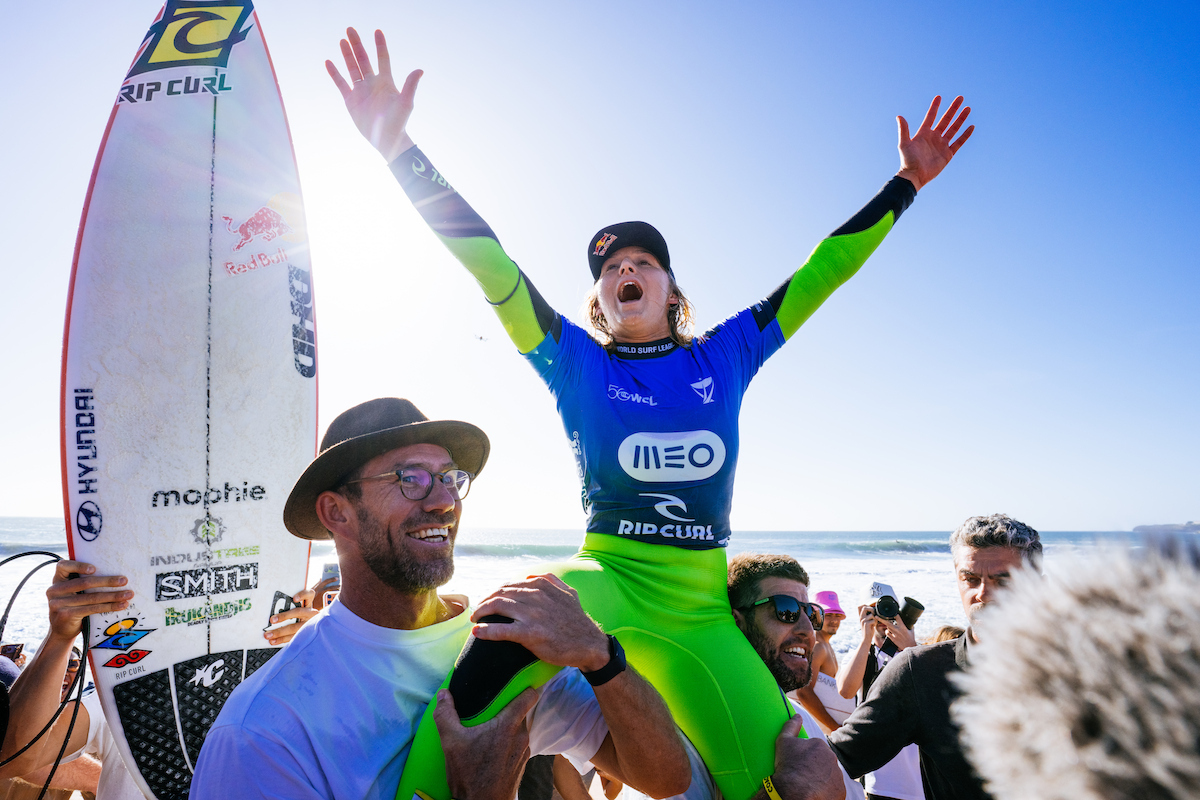 Molly Picklum in a blue jersey and green wetsuit with both arms raised, carried up the beach on a man's shoulders after her win