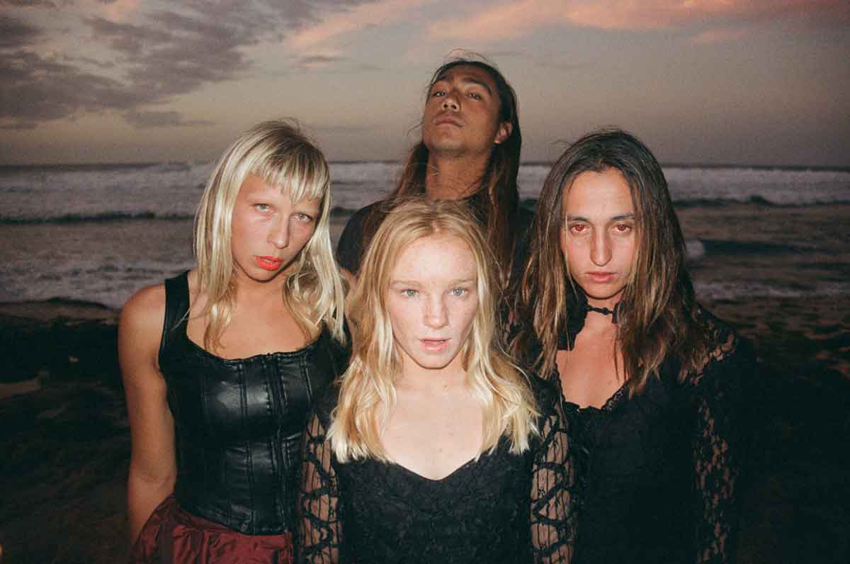 Four of the cast in black leather and lace, three with red eyes, stand close together on a beach at dusk with waves breaking behind