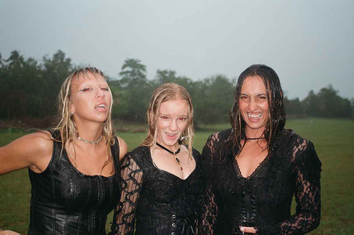 Three women in black corsets and lace, wearing red contact lenses, laugh in heavy rain on a grassy field