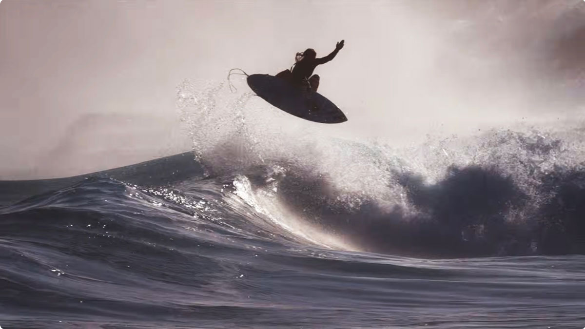 A surfer in a hooded wetsuit flies through the air above a dark breaking wave