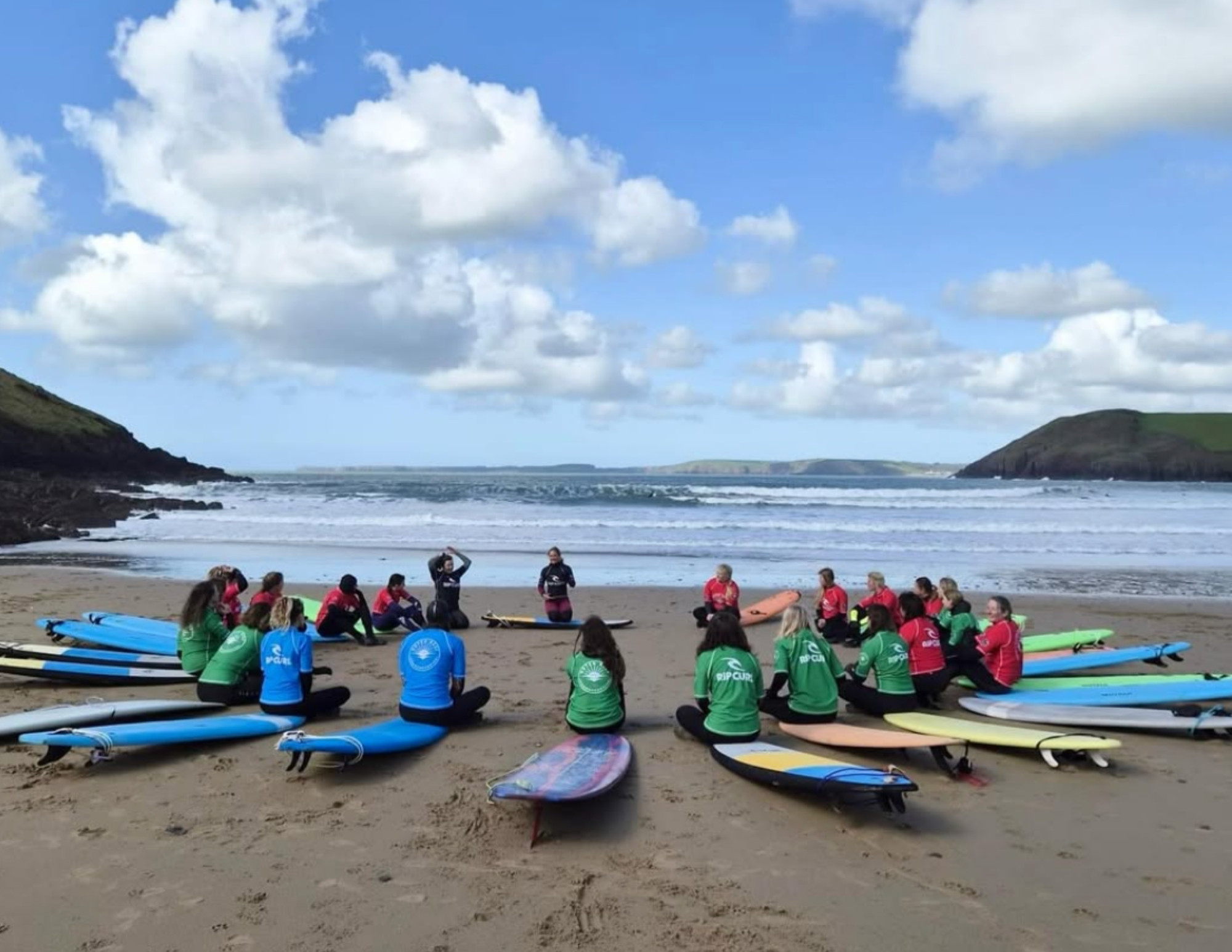 A surf school group sits in a circle on the beach with boards laid out around them, listening to an instructor