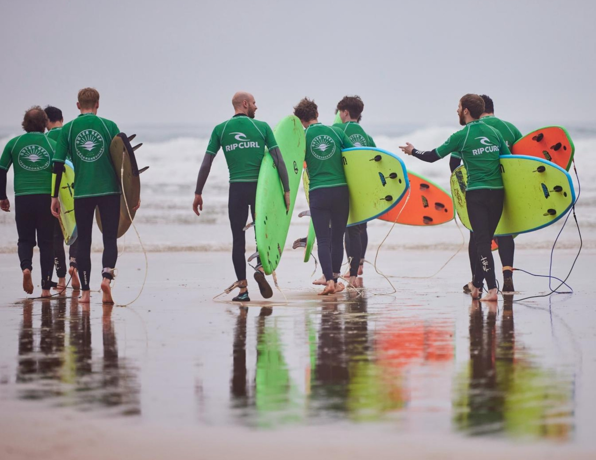 Surfers in green Outer Reef rash vests walk across wet sand towards the sea carrying foam boards