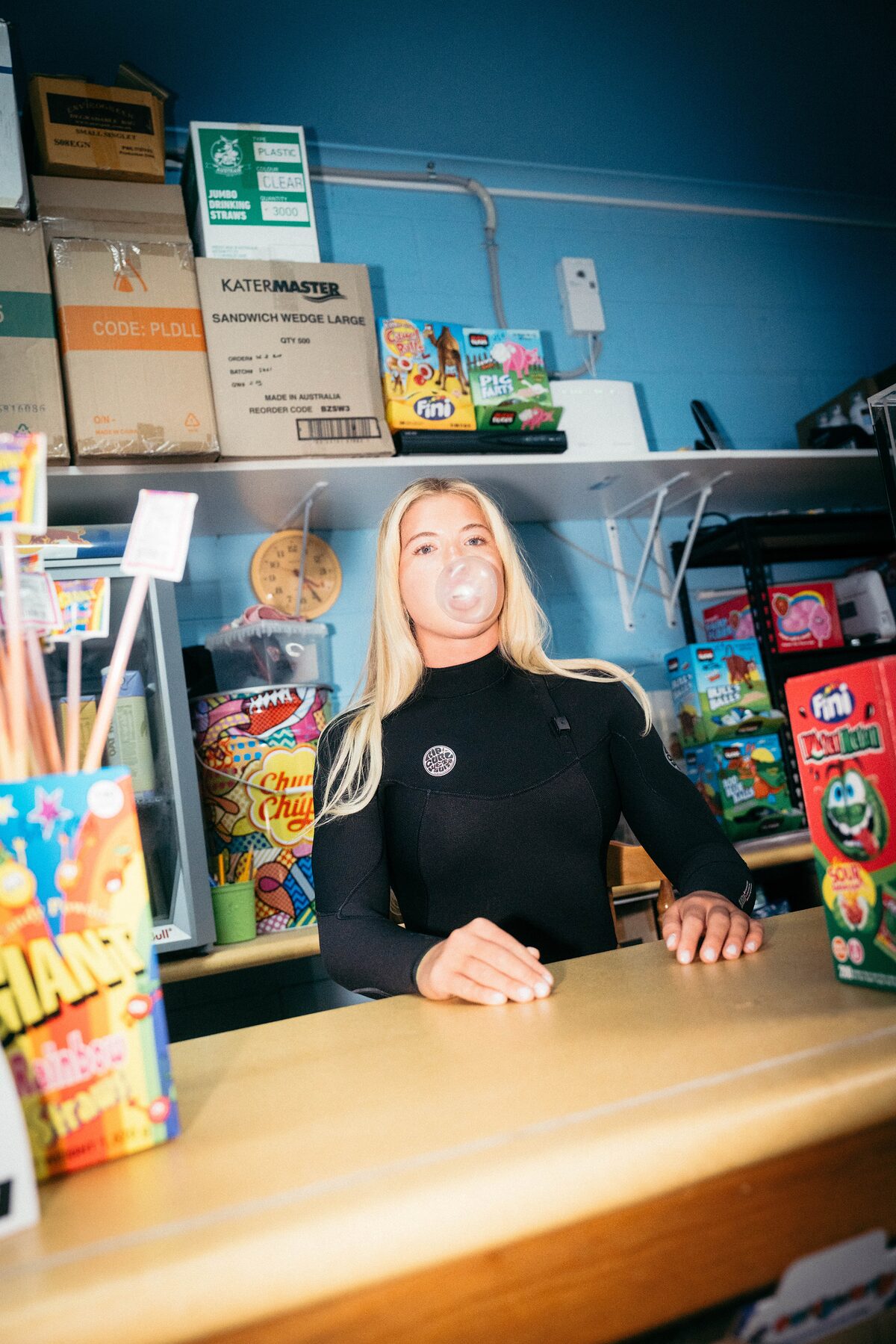 Woman in a wetsuit blowing a bubblegum bubble behind a shop counter