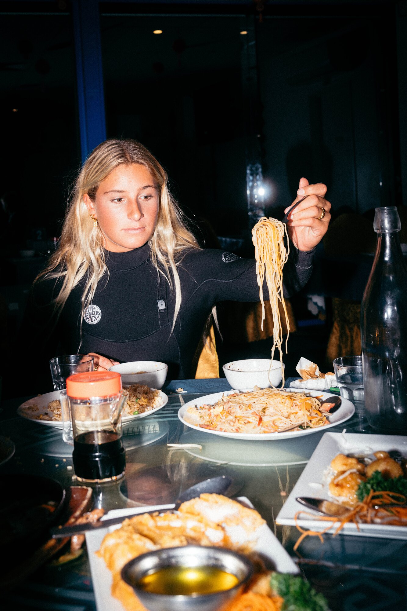 Surfer in a wetsuit eating noodles at a restaurant table