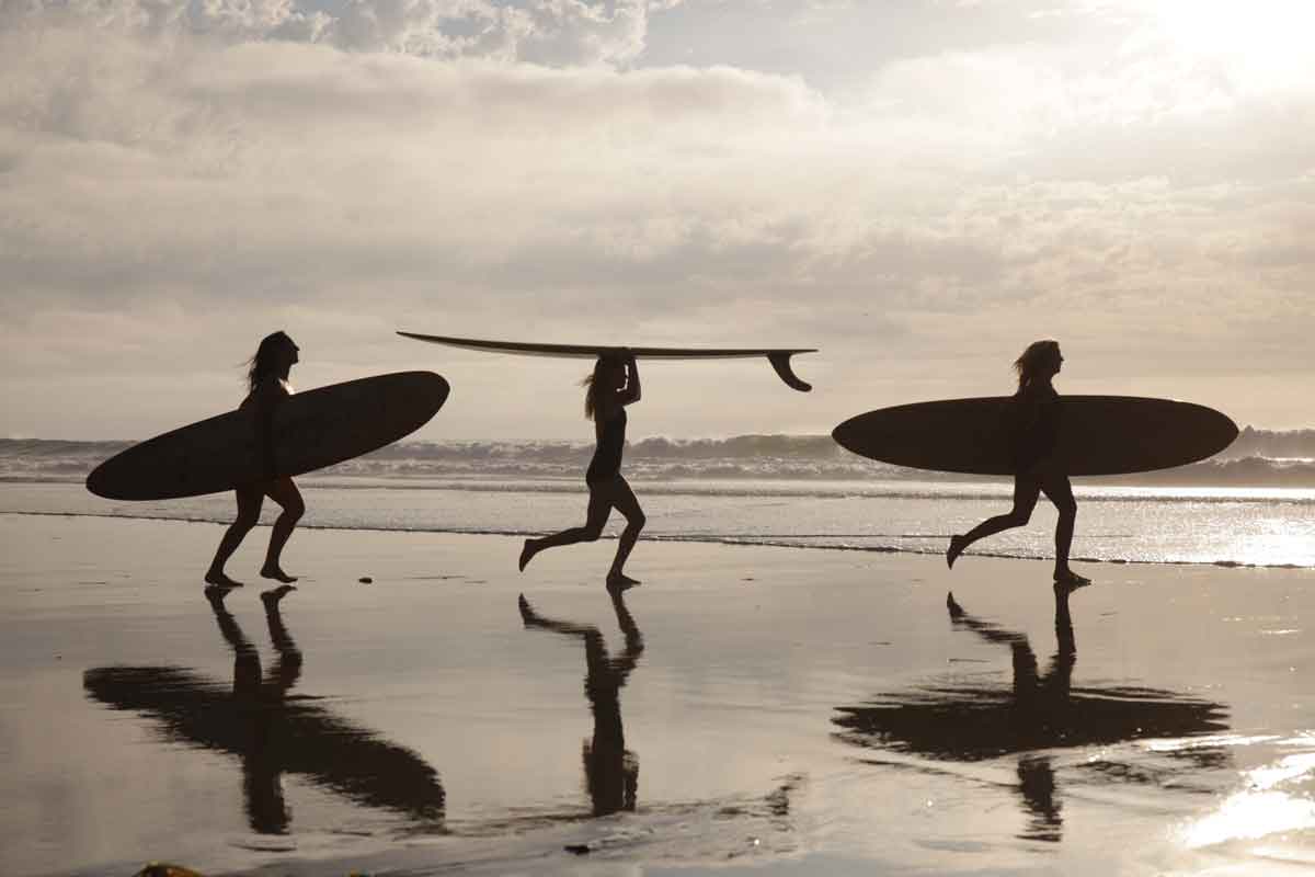 Three women in silhouette run along wet sand carrying longboards at sunset, their reflections in the shoreline