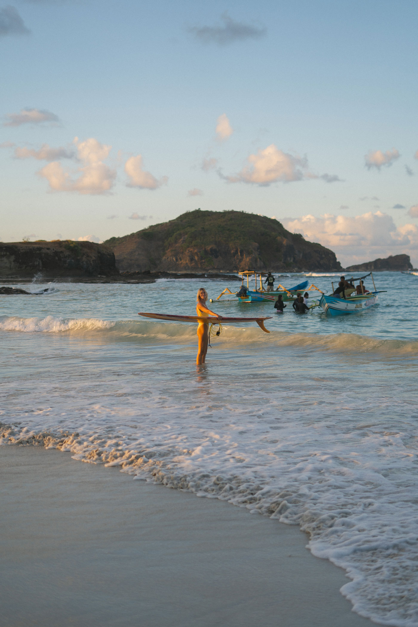 A woman in a yellow swimsuit stands in the shallows holding a single-fin surfboard, with outrigger boats and a headland behind at sunset